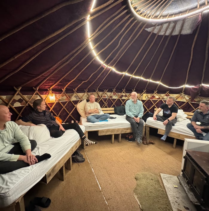 Photo of mens group with six men taking place in a yurt with wood stove
