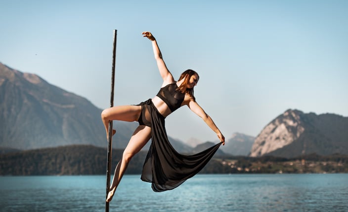 Sabrina Bel Pole doing a cupid pole trick outdoors, with a lake and mountains in the background.
