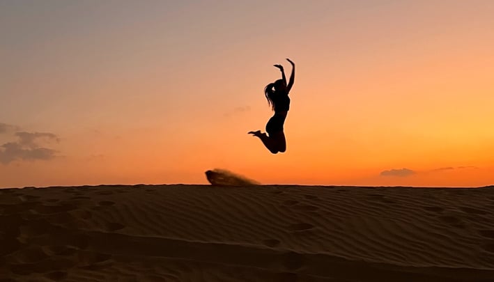 An image of a lady jumping over the sand dunes with a beautiful sunset on the background