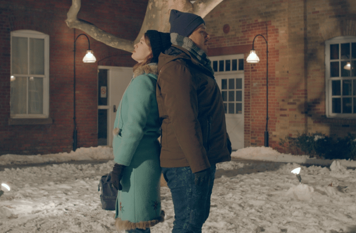 A young woman and man in winter clothing lean their backs against each other in a snowy courtyard