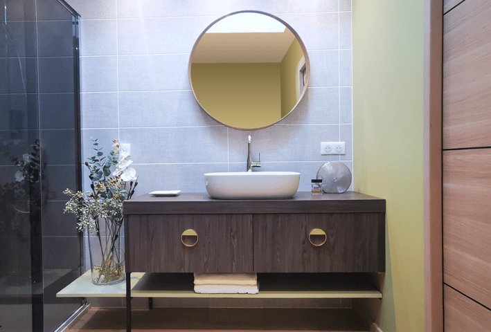 Salle de bains avec meuble en bois, poignées déco, tablette et miroir rond. vasque posée sur plan.
