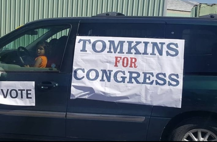 Banner with Tomkins for Congress on the side of a green minivan. A child can be seen in the window.