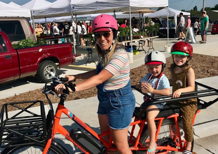 Woman stopped on a bike, posing with two young children seated in the back