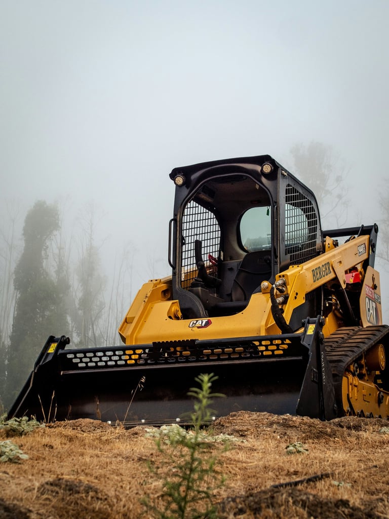 A yellow Caterpillar compact track loader parked on a foggy construction site with a front bucket attachment.