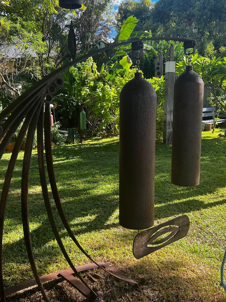 Rustic metal outdoor wind chimes hanging in a sunny tropical garden with lush green foliage.