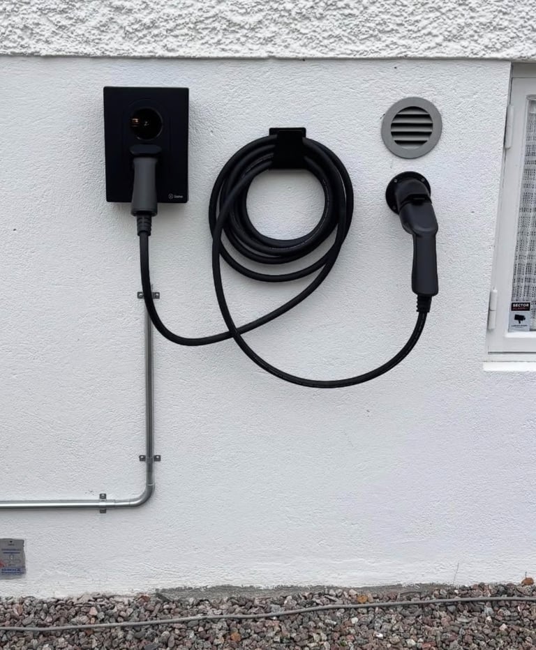 A wall-mounted black EV home charging station with a coiled cable and connector on a white exterior wall.