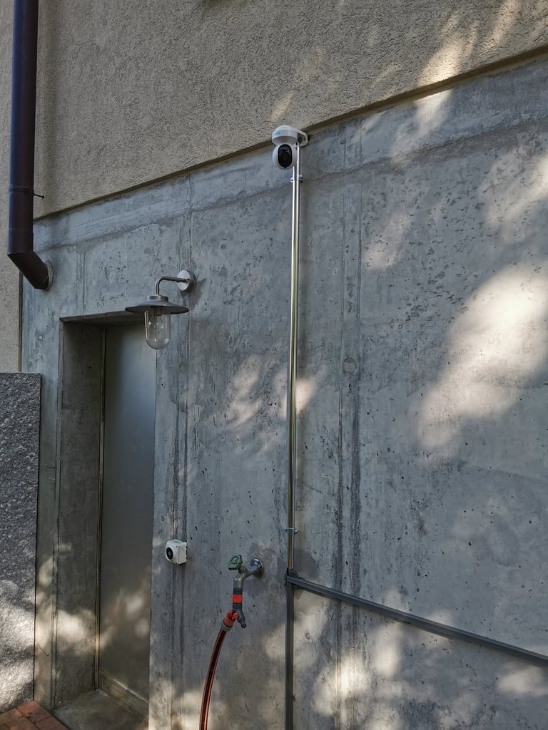 A white outdoor security camera mounted on a concrete wall with metal conduit and a wall lantern.