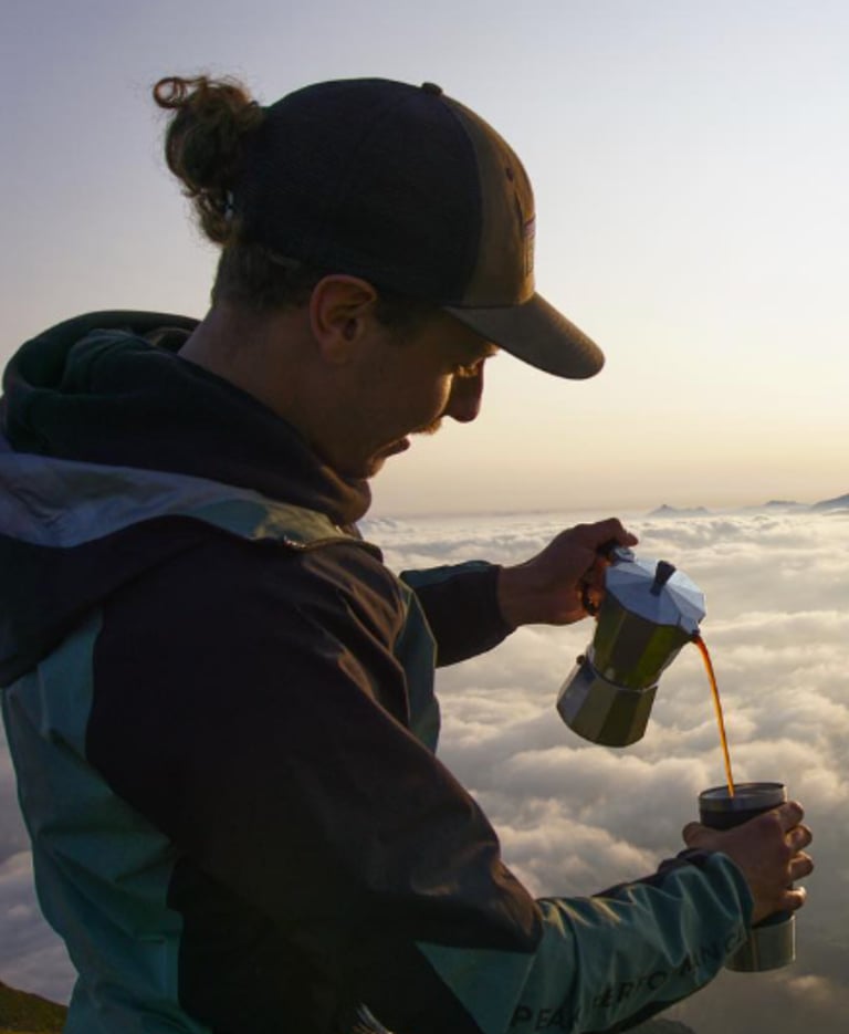 A man pours coffee from a moka pot into a mug while standing above the clouds during a mountain sunrise.