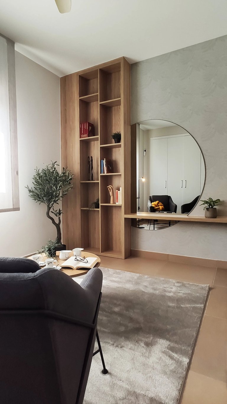 Interiorismo zona lectura con butaca gris, librería de madera y espejo redondo en San Antonio de Benagéber