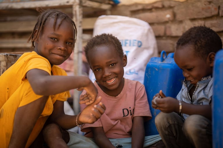Three Sudanese children smiling and playing near water containers.