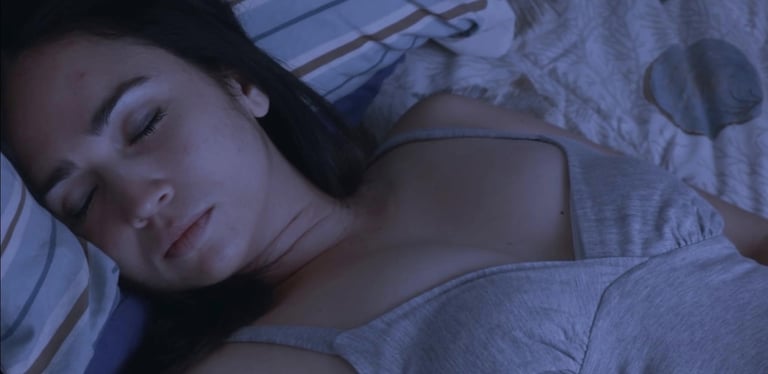 A dark-haired woman wearing a thin grey tank top is asleep in bed