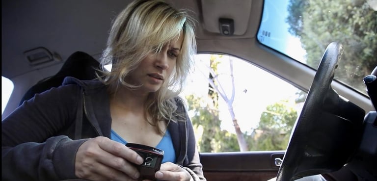 A young woman is sitting in the driver's seat of a stationary car with a mobile phone in her hand