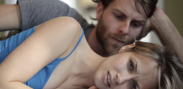 A bearded 30-something man lies on a bed looking down on the 30-something woman lying beside him. Neither one looks happy.