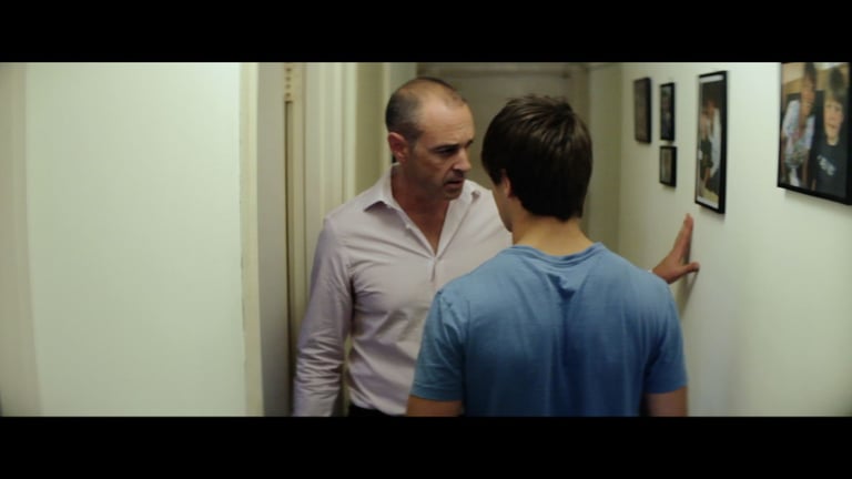 A young man faces an older man in a tense scene conducted in a narrow hallway space