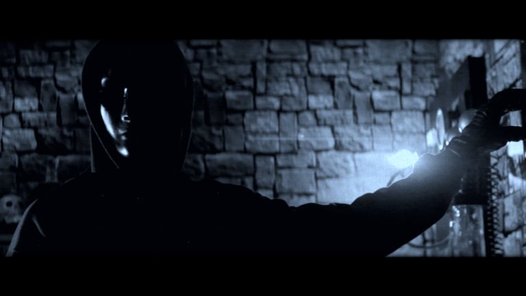 A man in a hooded sweater stands in a dimly lit room with brick walls with his outstretched arm poised to flick a wall switch