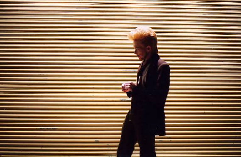 A young man with a bouffant hairdo and in a brown suit standing in front of a garage door with horizontal ridges