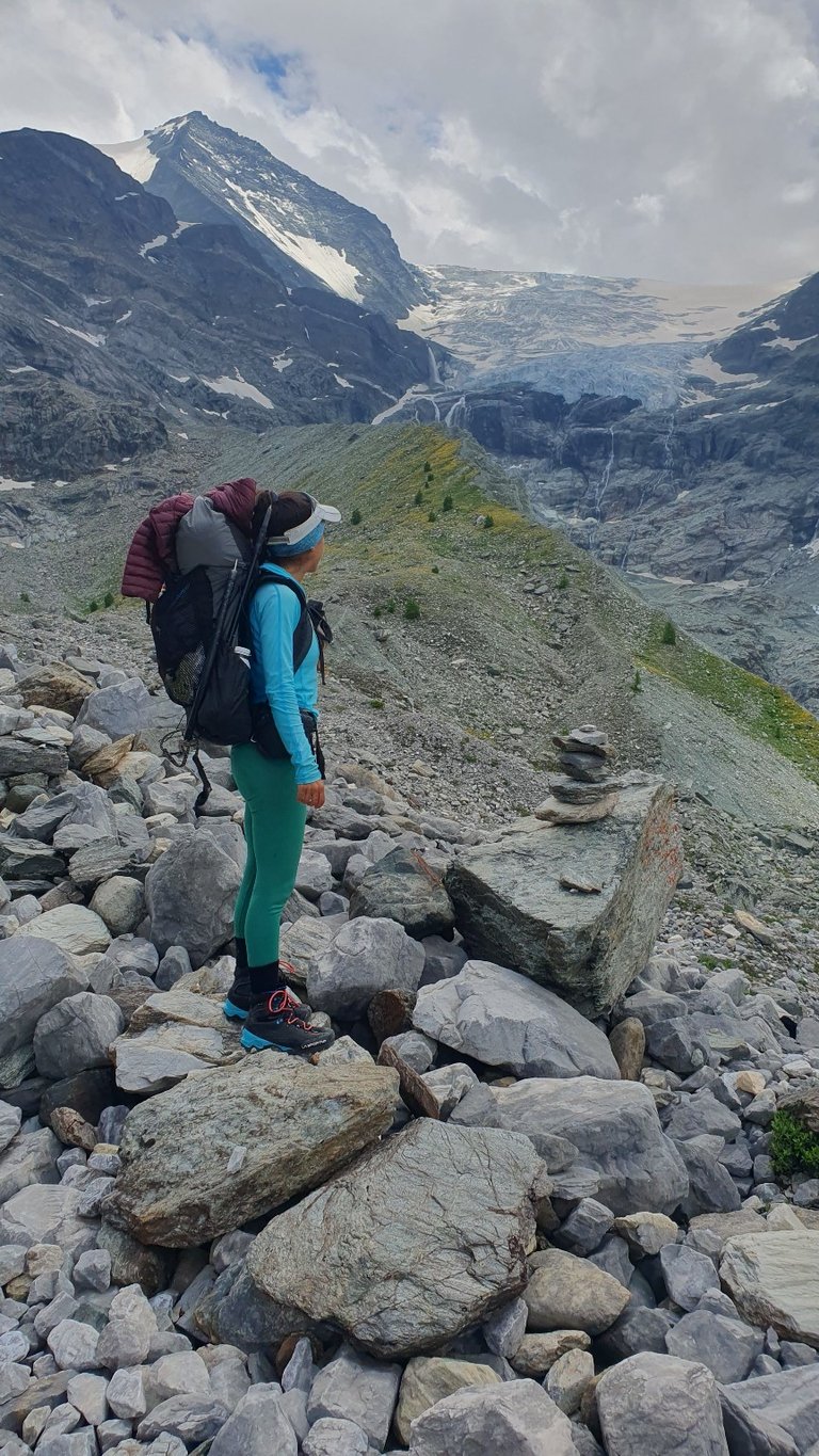 Female hiker with a backpack looking at a glacier and snowy mountain peaks on a rocky trail.
