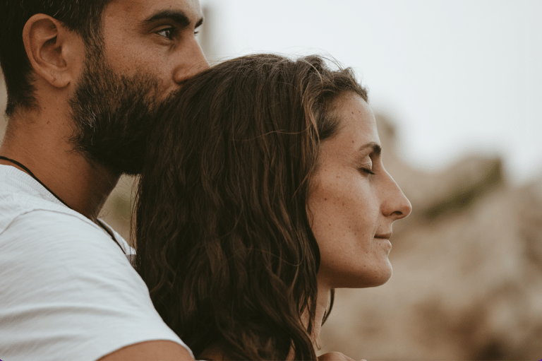 A bearded man kisses a woman with wavy hair on the forehead during a romantic outdoor sunset session.