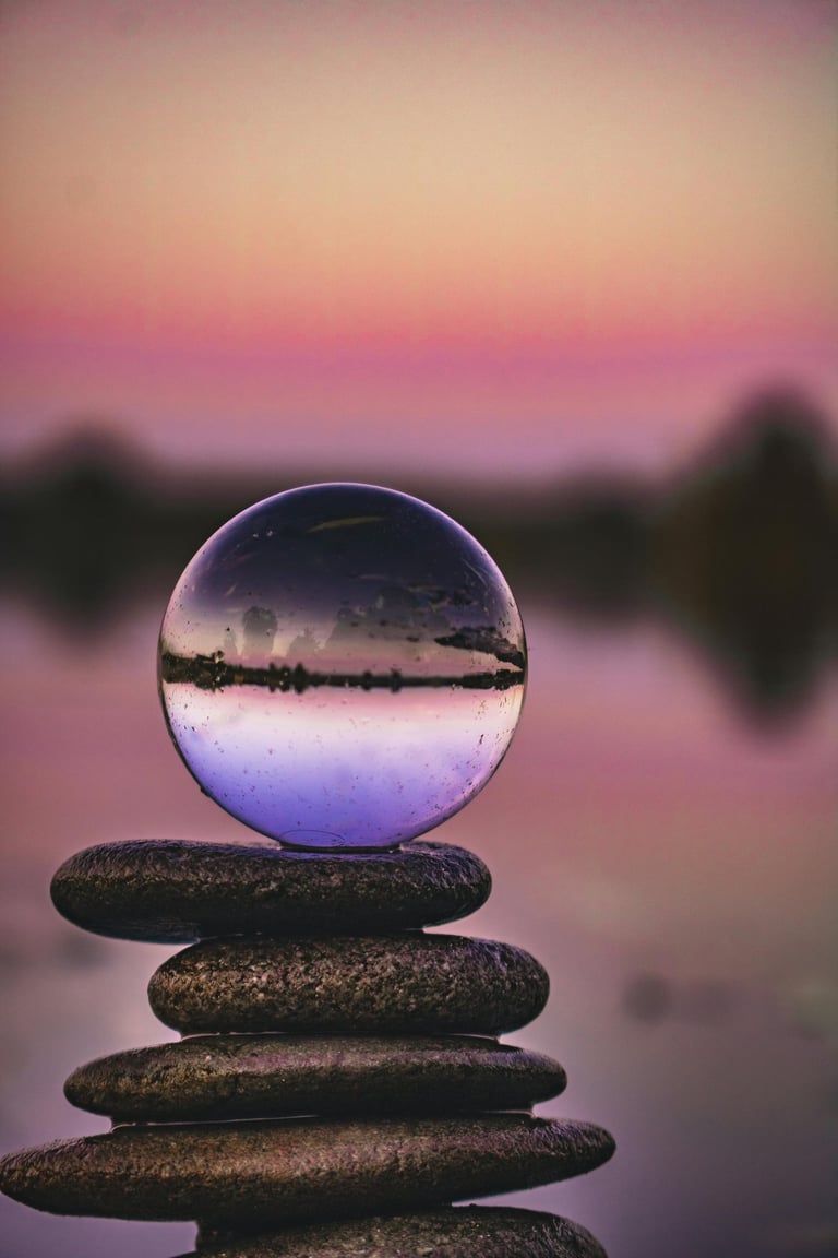 Crystal ball lensball balanced on stacked stones with a purple sunset reflection.