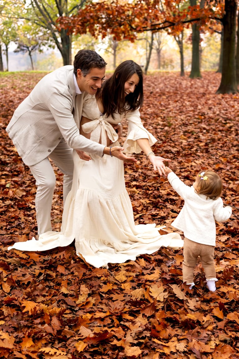 Smiling parents in autumn leaves holding hands with their toddler during an outdoor photo session.