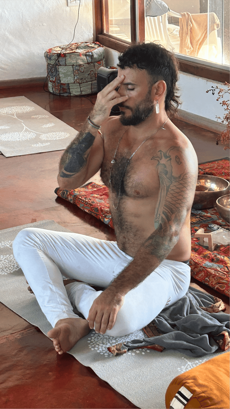 Tattooed man practicing alternate nostril breathing yoga on a mat in a sunlight studio.