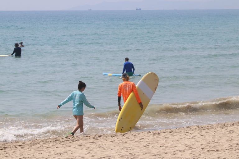A surf instructor in an orange shirt helps a student carry a yellow surfboard into the tropical blue ocean.