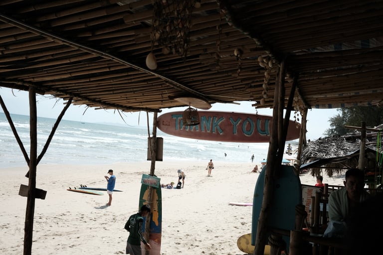Tropical beach scene viewed from a rustic bamboo surf shack with people on the sandy shore and ocean waves.