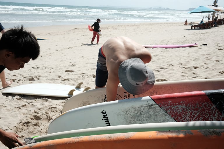 Surfers organizing surfboards on a sunny tropical beach with ocean waves in the background.
