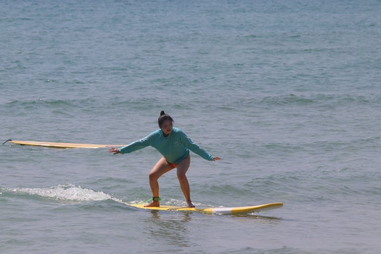 A female surfer in a blue long-sleeve rash guard balances on a yellow surfboard riding a small ocean wave.