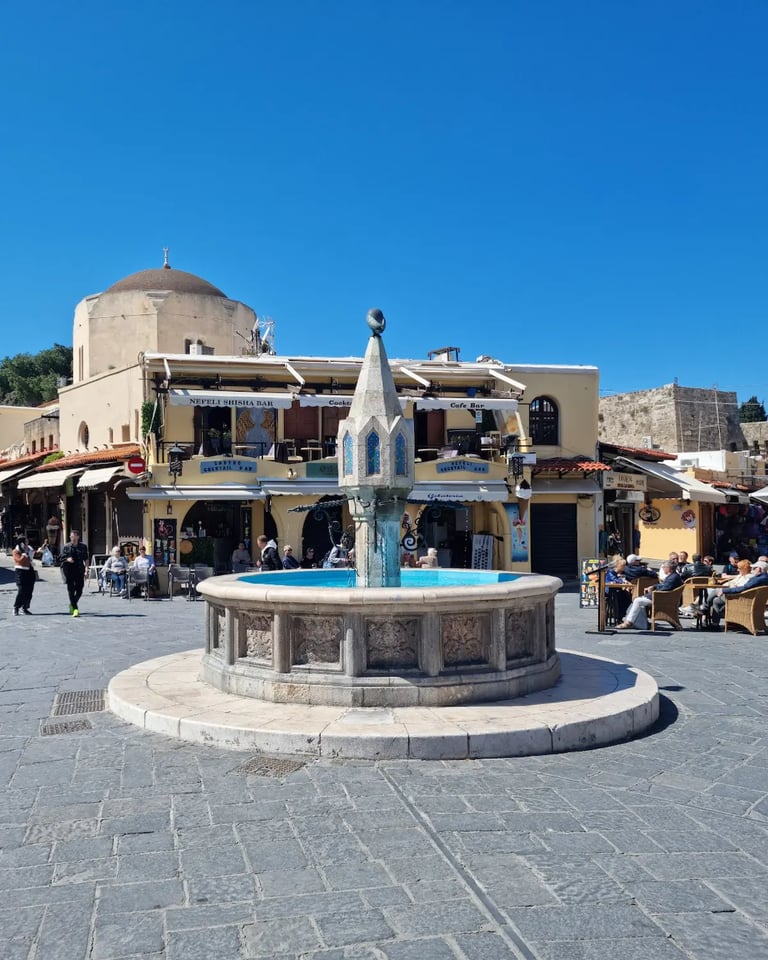 The ornate stone fountain in Hippocrates Square, Rhodes Old Town, with outdoor cafes and a mosque under a blue sky.