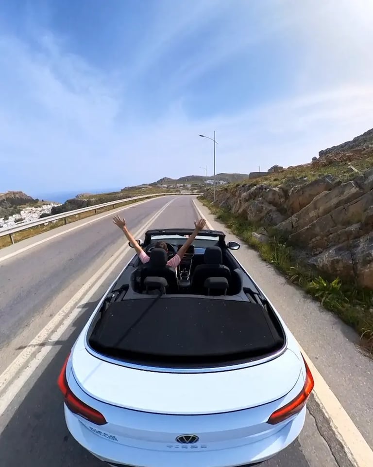 A driver with arms raised in a white Volkswagen T-Roc convertible driving on a coastal road.