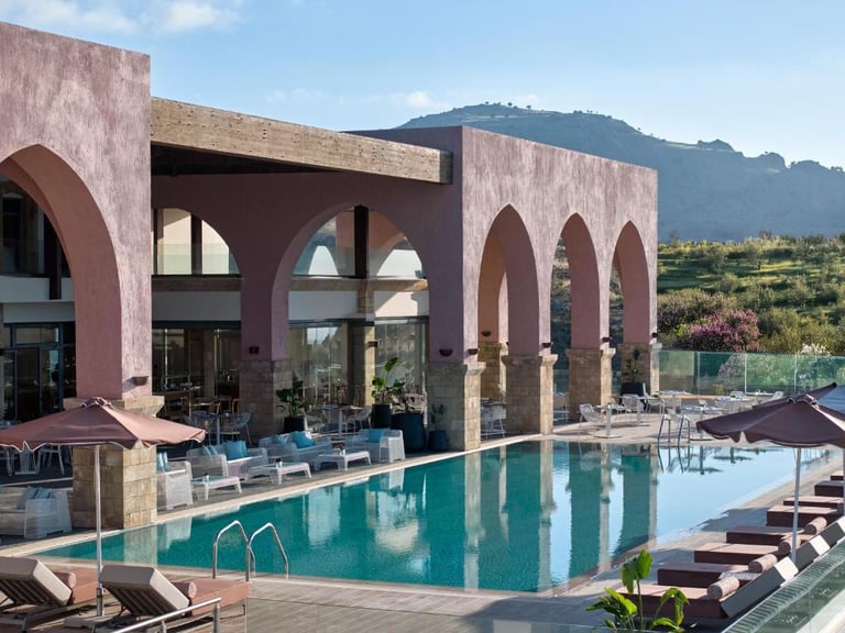 Luxury resort swimming pool with pink arched architecture and sun loungers under a mountain backdrop.