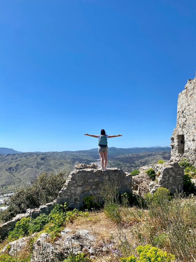 A hiker with outstretched arms stands on stone ruins overlooking a vast mountain valley under a clear blue sky.