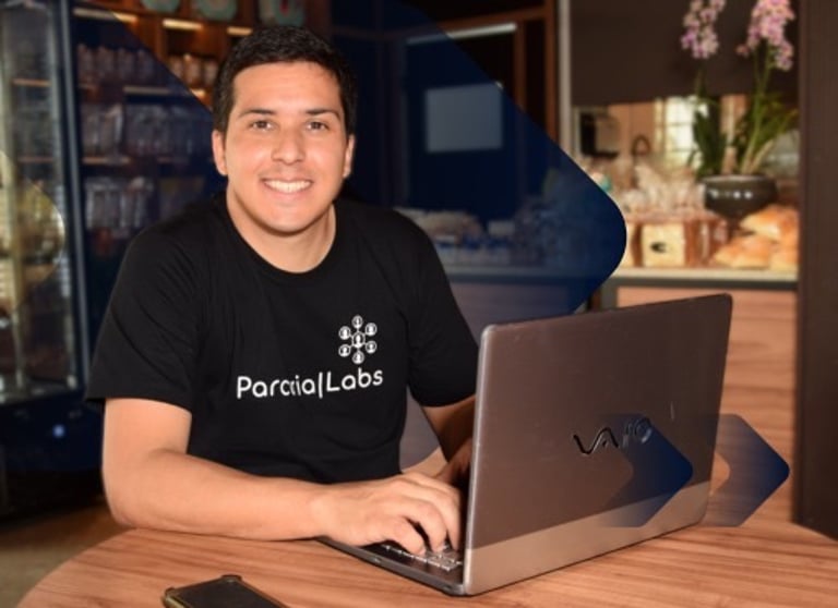 Douglas Silva, homem sorrindo, com camiseta preta, em uma cafeteria, sentado com seu notebook