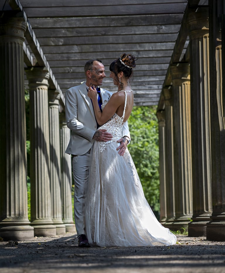 Ein Hochzeitspaar blickt sich verliebt an, während es unter einer Holzpergola mit Steinsäulen steht; die Braut trägt ein träg