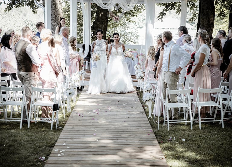 Dieses Farbfoto zeigt zwei Bräute, die in ihren Hochzeitskleidern einen Holzsteg entlanggehen, der mit verstreuten Blütenblät