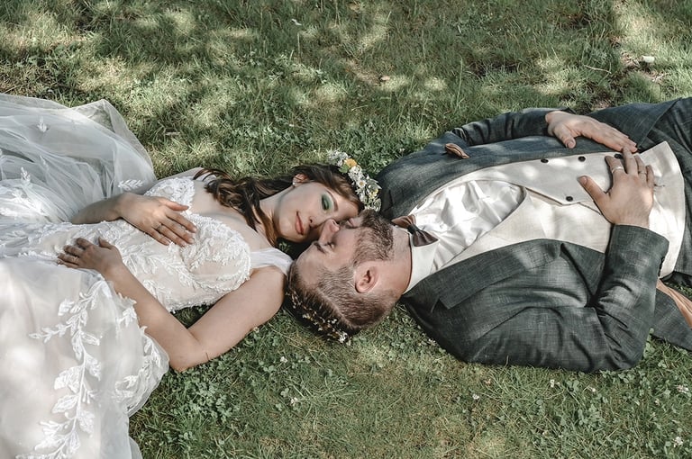 Dieses Farbfoto zeigt ein Brautpaar, das auf grünem Gras liegt und sich ansieht. Die Braut trägt ein weißes Hochzeitskleid mi