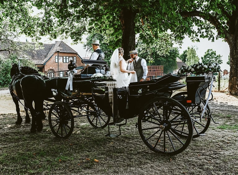 Dieses Farbfoto zeigt eine Hochzeitskutsche mit Pferden und drei Personen unter großen, belaubten Bäumen. Eine Braut in einem