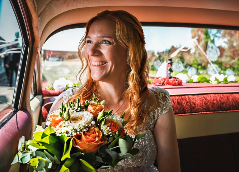 Glückliche Braut mit Blumenstrauß sitzt im Oldtimer und lächelt aus dem Fenster, Sonnenlicht fällt auf ihr Gesicht.