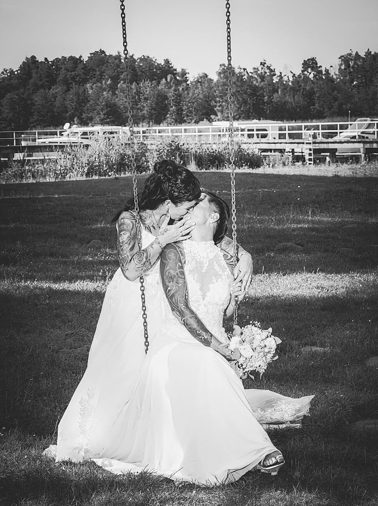  Dieses Schwarz-Weiß-Bild zeigt zwei Bräute in weißen Hochzeitskleidern, die sich küssen, während sie auf einer Schaukel sitz