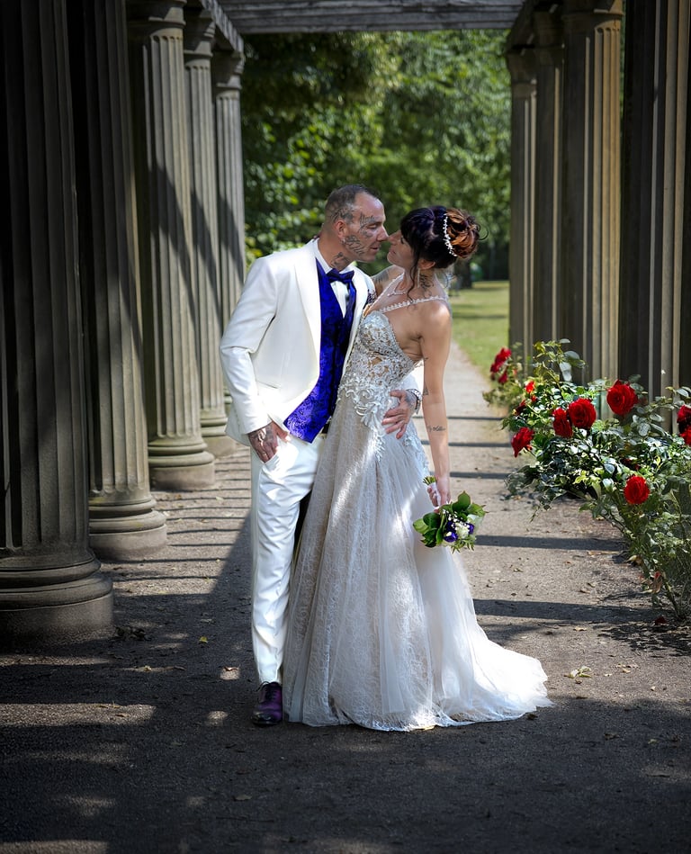 Ein Hochzeitspaar küsst sich unter einer Pergola mit Steinsäulen und einer Reihe roter Rosen; der Bräutigam trägt einen weiße