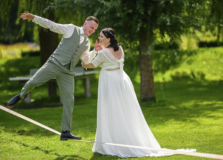 Dieses Farbfoto zeigt ein Brautpaar in einer spielerischen Pose auf einer Slackline in einem grünen Park. Der Bräutigam, gekl