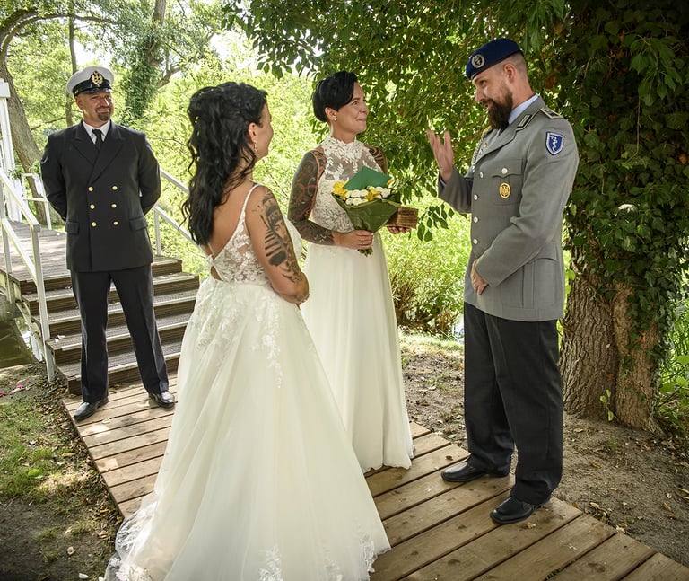 wei Bräute und zwei Männer in Militäruniformen unter einem Baum auf einem Holzsteg, eine Braut hält einen Blumenstrauß.