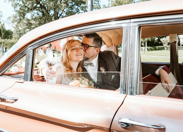 Braut und Bräutigam sitzen glücklich in einem Oldtimer, der Bräutigam küsst die Braut liebevoll auf die Wange, Brautstrauß im