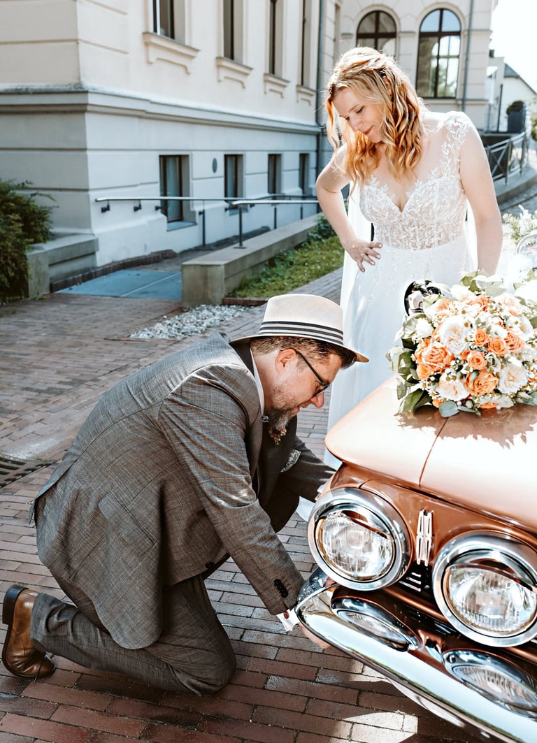Bräutigam kniet vor einem Oldtimer und prüft das Auto, während die Braut in einem weißen Spitzenkleid lächelnd zusieht, Hochz