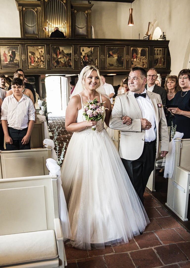  Ein Bild, das eine lächelnde Braut zeigt, die von einem Mann, wahrscheinlich ihrem Vater, den Gang einer Kirche entlanggefüh