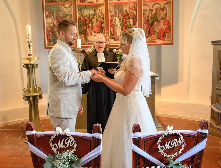 Dieses Farbfoto zeigt eine Hochzeitszeremonie in einer Kirche. Im Mittelpunkt stehen Braut und Bräutigam, die sich gegenseiti
