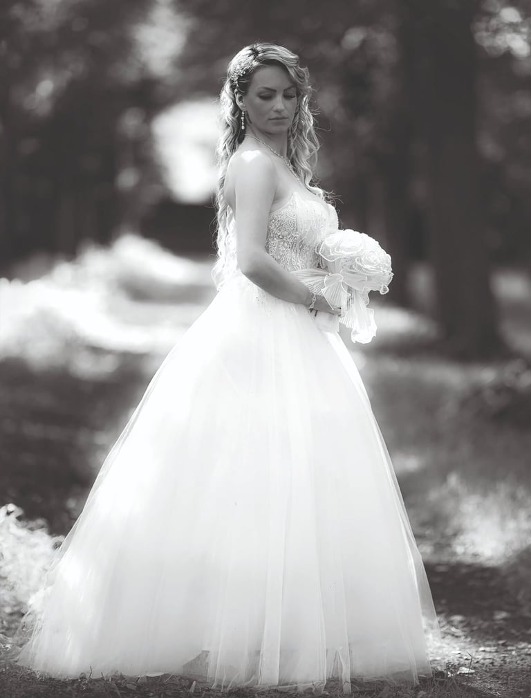 Braut im eleganten weißen Hochzeitskleid steht im Wald und hält ihren Blumenstrauß – romantisches Schwarz-Weiß-Hochzeitsfoto 