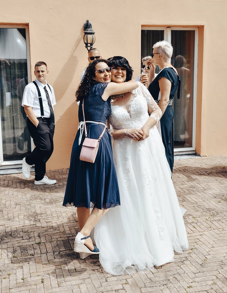 Eine Braut in einem weißen Hochzeitskleid wird von einer Freundin in einem dunkelblauen Kleid umarmt, während andere Hochzeit
