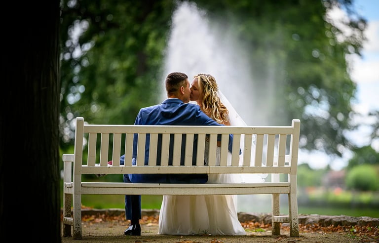 Ein Brautpaar sitzt auf einer weißen Bank in einem Park und küsst sich innig, während im Hintergrund ein großer Springbrunnen